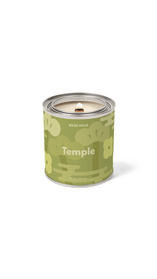 A single lit candle in a cylindrical metal tin with a green floral label reading 'Temple' and 'BASK HOUR'. The candle is shown on a clean white background with a soft shadow.