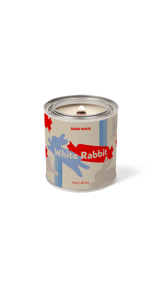 A single candle in a cylindrical tin with a lid, featuring a label that reads 'White Rabbit' and 'BASK HOUR.' The label has a beige base with a blue rabbit silhouette, red abstract shapes, and blue vertical stripes.