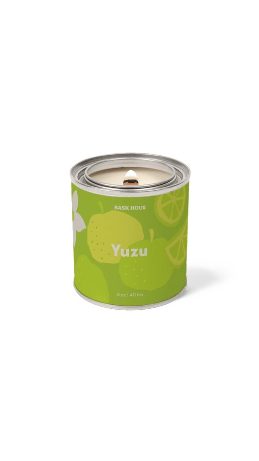 A single cylindrical scented candle in a metallic tin with a bright green label reading 'Yuzu' and 'Bask Hour.' The label includes yellow citrus illustrations and a white flower graphic.